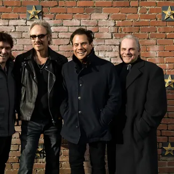 Five men stand in a row, posing and smiling in front of a brick wall decorated with gold stars.