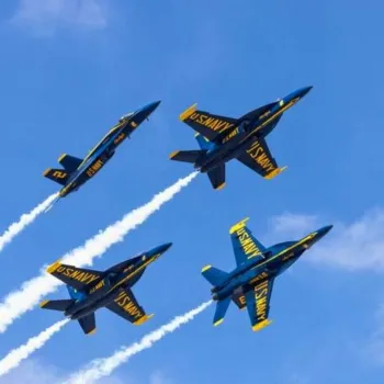 Four U.S. Navy Blue Angels jets fly in formation against a clear blue sky, leaving white trails behind them.