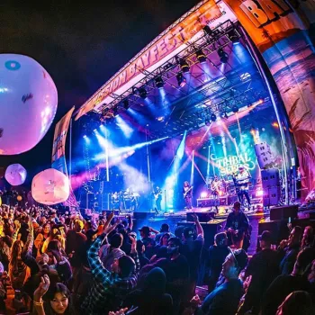 Large crowd at an outdoor music festival at night, with giant balloons in the air and a band performing on a brightly lit stage.