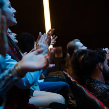 Audience members sit in theater seats and clap, facing forward, in a dimly lit environment with a vertical light in the background.