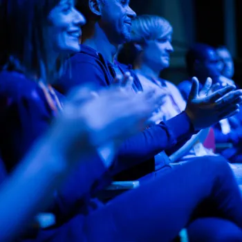 A group of people sits in a row, clapping and smiling, bathed in blue stage lighting at what appears to be an indoor event or performance.