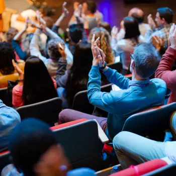 Audience members seated in rows clap and watch a person on stage during an indoor event or presentation.