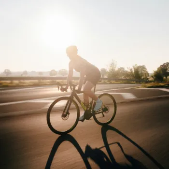 A cyclist rides a road bike on an empty road during sunrise or sunset, casting a long shadow. Trees and open fields can be seen in the background.