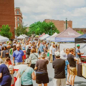 A large crowd of people walk among vendor tents and tables at an outdoor street market on a sunny day, with brick buildings and trees in the background.