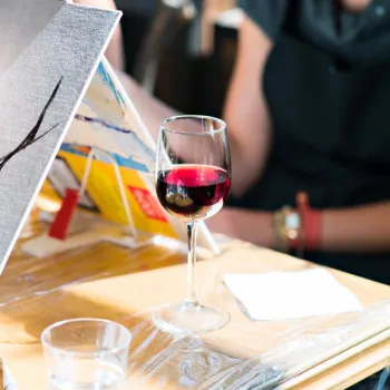 A person paints on a canvas with a brush at a table, next to a glass of red wine, a glass of water, and another painter in the background.