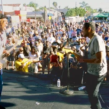 Two musicians play electric guitars on an outdoor stage facing a large, attentive crowd during a daytime event.