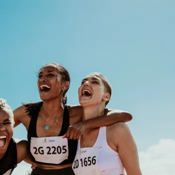 Three women in athletic wear and race bibs smile and laugh together under a bright blue sky after a race.