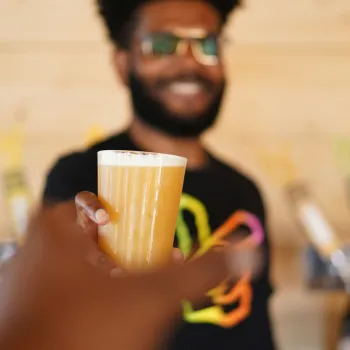 A man with a beard and glasses holds out a glass of craft beer, with several colorful tap handles in the background.