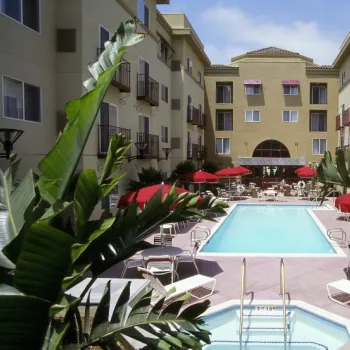 A hotel courtyard features a central outdoor swimming pool surrounded by lounge chairs, tables with red umbrellas, and tall plants in the foreground.