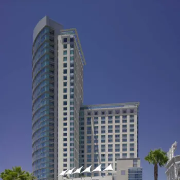 A modern high-rise hotel building with glass and beige exterior stands under a clear blue sky, surrounded by palm trees and a street with blurred moving vehicles in front.