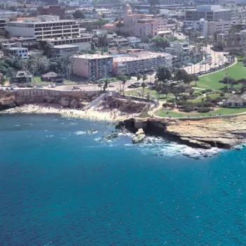 A coastal city scene with parks, rocky shoreline, buildings, and clear blue water extending into the distance.