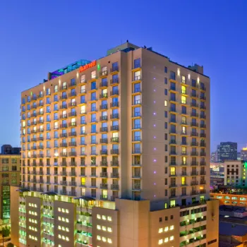 A tall, modern Marriott hotel building is lit up at dusk in an urban setting, with cars parked along the surrounding streets.