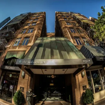 Wide-angle view of the entrance to The Sofia Hotel, a brick building with green canopies, located at 150 W Broadway on a sunny day.