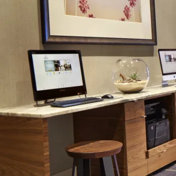 Two desktop computers on a wooden counter with stools, a glass bowl with plants in the center, and a framed artwork on the wall behind.
