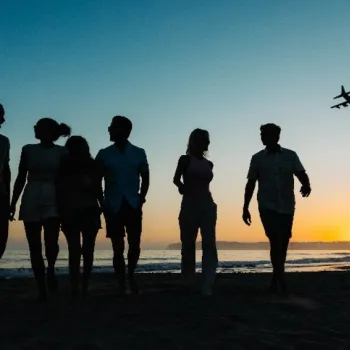 Six people walk along a beach at sunset with an airplane flying in the sky above the horizon.