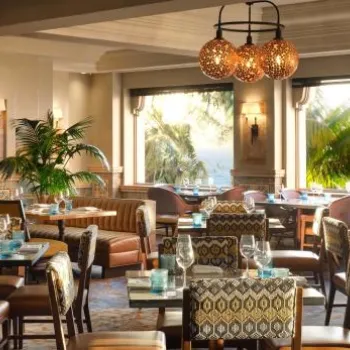 Elegant restaurant interior with patterned chairs, set tables, a decorative fireplace, large windows, palm trees, and an ocean view at sunset.