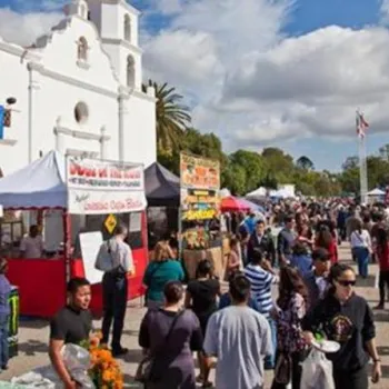 Crowds attend an outdoor Día de los Muertos festival with food stalls and decorations near a white mission-style building under a cloudy sky.