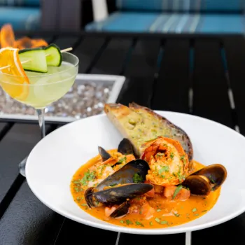 A white bowl of seafood stew with mussels and shrimp in a red sauce, served with grilled bread, sits next to a cocktail garnished with orange and cucumber slices on an outdoor table.