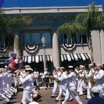 A marching band in white uniforms performs in a parade on a sunny day, with a crowd watching and American flags and bunting decorating buildings in the background.