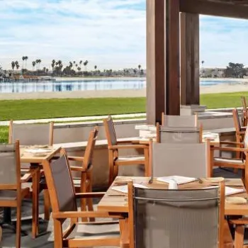 Outdoor restaurant seating area with wooden tables and chairs overlooking a grassy lawn and a calm waterfront with sailboats in the background.