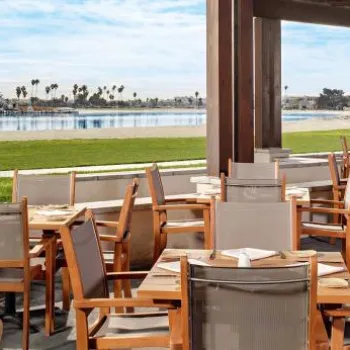 Outdoor restaurant seating area with wooden tables and chairs overlooking a grassy lawn and a calm waterfront with sailboats in the background.
