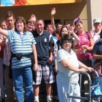 A group of people, some waving and smiling, pose for a photo outside a building. One person at the front uses a walker.