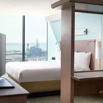 Modern hotel room with a bed, desk, office chair, flat-screen TV, and large window offering a view of a harbor and a docked military ship.