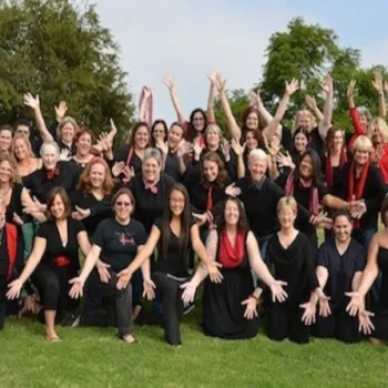 A large group of people dressed mostly in black with some red accents pose outdoors on grass, smiling and holding their arms out in a welcoming gesture.