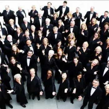 A large group of people dressed in formal black attire stand together, facing the camera and raising their hands in a wave.