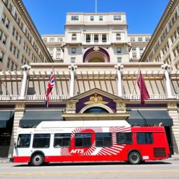 A red and white MTS city bus passes in front of a large, multi-story historic hotel with flags and arched entrances.