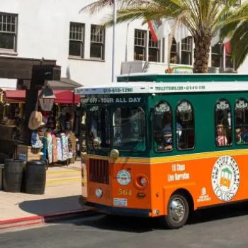 A green and orange trolley drives past shops and palm trees in a sunny outdoor shopping area with people standing nearby.