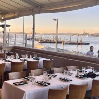 Outdoor restaurant patio with set tables overlooks a waterfront and city skyline; two people walk along the promenade in the background.