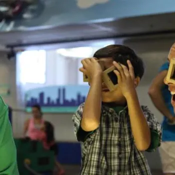 An older man, a young boy, and a younger man sit at a table indoors; the boy and man hold small objects to their eyes while the older man smiles.