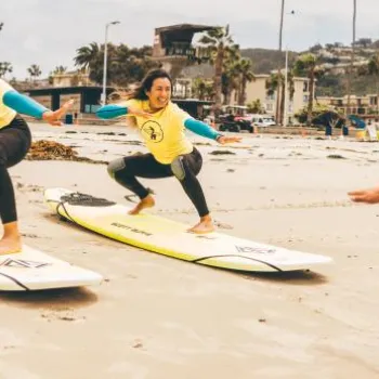 Two people in yellow shirts practice surfing stances on boards in the sand while an instructor in a red shirt coaches them on the beach.