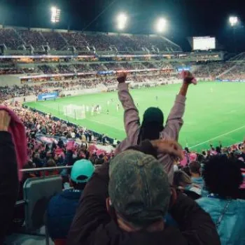 A large crowd of fans cheers during a night soccer match in a brightly lit stadium.