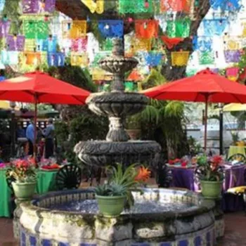 Outdoor courtyard with a central stone fountain, colorful tablecloths on tables, red and yellow umbrellas, and bright papel picado banners hanging overhead.