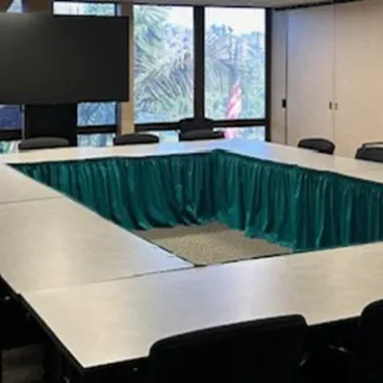 Conference room with rectangular table arrangement and black chairs, large windows showing trees outside, and a large screen at the front.
