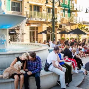 People sitting and socializing around a large fountain and outdoor café tables in a lively urban plaza with modern buildings in the background.