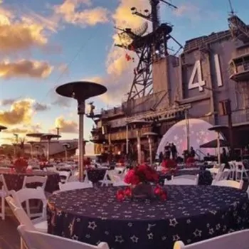 Tables with star-patterned tablecloths and chairs are set up for an event on the deck of an aircraft carrier at sunset, with the number 41 visible on the ship.