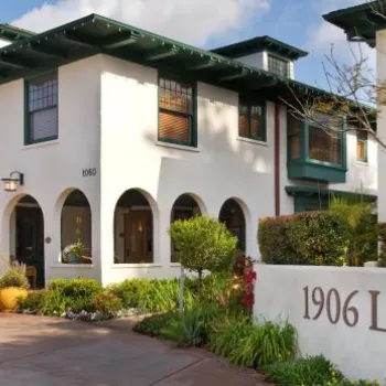 White two-story building with green trim and arched windows, landscaped garden, and a sign reading "1906 Lodge" near the entrance.