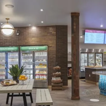 A modern café interior with refrigerated display cases, a counter with a blue tiled front, digital menu screens, and wooden tables with benches.