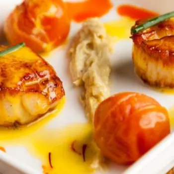 Seared scallops and roasted cherry tomatoes on a white plate, garnished with sauce and a vegetable puree.