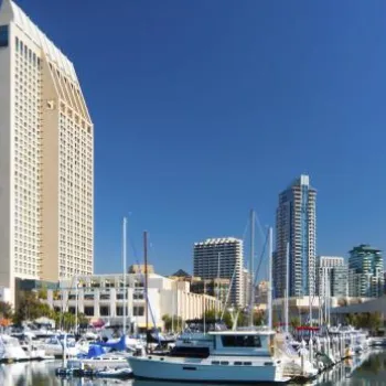 Marina with docked boats in the foreground and modern high-rise buildings under a clear blue sky in the background.