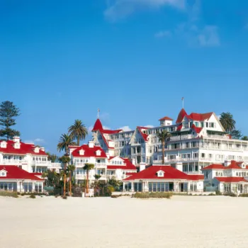 A large beachfront hotel with distinctive red roofs and white walls is set against a blue sky, with sand and palm trees in the foreground.