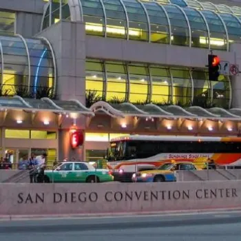 People stand outside the San Diego Convention Center, with buses and cars parked in front and the building’s glass facade visible in the background.