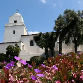 White mission-style building with a bell tower, surrounded by trees and colorful flowering plants under a partly cloudy sky.