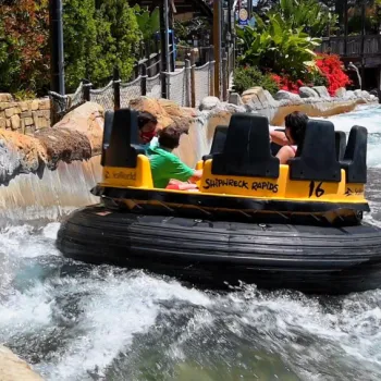 Four people ride a circular yellow raft labeled "Shipwreck Rapids" along a water channel surrounded by rocks and greenery at an amusement park.