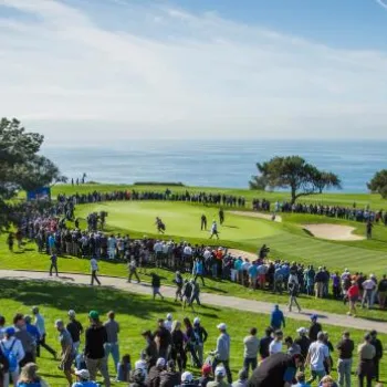 A large crowd watches golfers compete on a green near the ocean, with clear skies and trees surrounding the course.