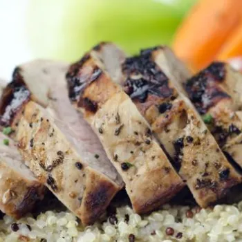 Sliced grilled meat served on a bed of quinoa, garnished with a sprig of rosemary, with carrots and greens in the background.