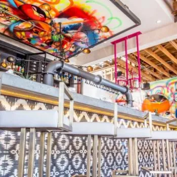 A modern bar with high stools, colorful abstract graffiti art on the walls and ceiling, exposed wooden beams, and geometric patterned tiles.